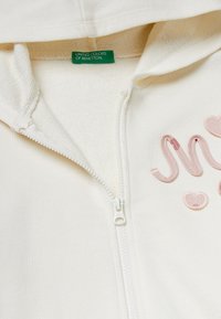 Felpa zip-up color crema con dettagli in rosa ricamati. Presenta una texture morbida, cappuccio e un'etichetta verde con la scritta "UNITE COLORS OF BENETTON."