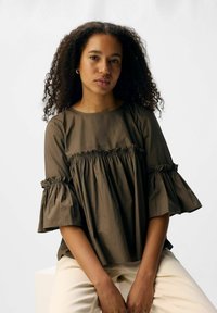 Olivenfarvet bluse med flæser på ærmerne og smock-detaljer. Blødt, glat stof med en afslappet pasform. Kombineret med lyse bukser.