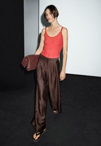 Rode geribbelde tanktop en losse bruine geplooide broek. Model houdt een gestructureerde bordeauxrode tas vast. Geplaatst tegen een minimalistische achtergrond.