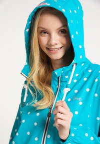 Jacket de ploaie turcoaz cu buline albe, cu glugă, având un sistem de strângere cu șnur și un etichetă inscripționată cu "love". Material neted, rezistent la apă.