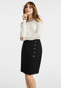 Czarna spódnica ołówkowa z złotymi guzikami po bokach i białą bluzką z długim rękawem z czarną lamówką przy dekolcie. Gładka tekstura materiału.