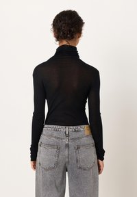 Keeruliste juustega inimene, kes kannab keha järgivat musta pikkade varrukatega pluusi ja kõrge vöökohaga halli teksapükse, seismas valge ühevärvilise tausta ees.