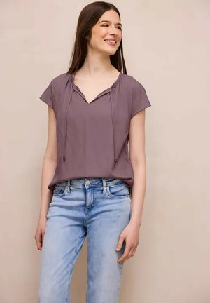 Młoda kobieta uśmiecha się, ubrana w fioletową bluzkę z krótkim rękawem i wiązaniami oraz jasnoniebieskie dżinsy, stoi na tle jednolitego beżowego tła.