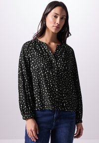 Schwarze Bluse mit einem strukturierten Muster aus verstreuten weißen Formen, mit einem geknöpften V-Ausschnitt und gerafften Ärmeln. Kombiniert mit blauen Jeans.