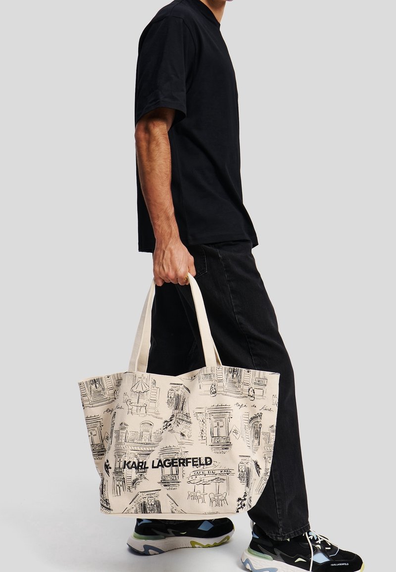 Personne en tenue noire tenant un sac fourre-tout beige avec des croquis architecturaux noirs et le texte « KARL LAGERFELD », portant des baskets noires.