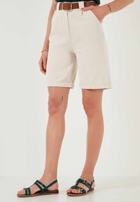 Pantaloni corti color crema, fino al ginocchio, con orli arrotolati, abbinati a una cintura marrone. I sandali presentano cinturini verde scuro e hardware minimalista.