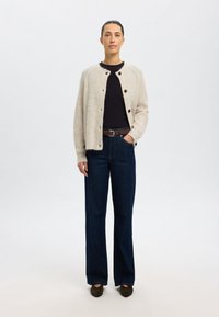 Beige gebreide cardigan met ronde hals en zwarte knopen, gedragen over een zwart topje. Gecombineerd met hooggepolijste blauwe denim jeans en donkere schoenen.