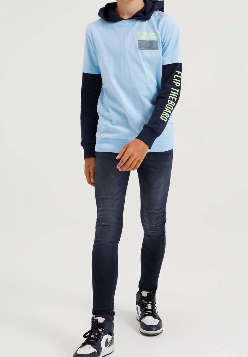 Person som bär en ljusblå hoodie med marinblå ärmar, tryckt text, mörka skinny jeans och svart-vita högklackade sneakers.