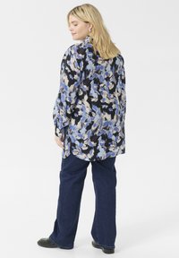 Blumenbluse in Schwarz, Blau und Creme, mit lockerer Passform und langen Ärmeln. Kombiniert mit dunkelblauen weit geschnittenen Jeans und schwarzen Schuhen.