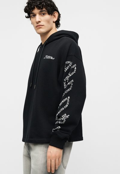 Jeune homme portant un sweat à capuche noir avec du texte blanc sur la poitrine et la manche, associé à un jean gris clair, debout devant un fond uni.