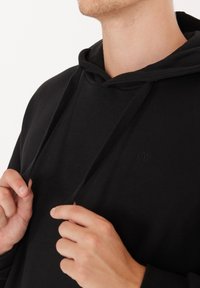 Henkilö säätää mustan hupparin nauhoja, näyttäen kaula-aukon ja pienen brodeeratun logon rinnassa.