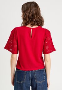 Haut rouge avec des manches courtes en dentelle et une fermeture en trou de serrure au dos. Tissu texturé avec une coupe légèrement ample, porté avec un jean taille haute.