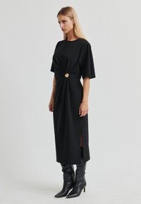 Robe midi noire à manches courtes, détail noué à la taille avec un bouton doré, fentes sur les côtés, assortie à des bottines noires à talons.