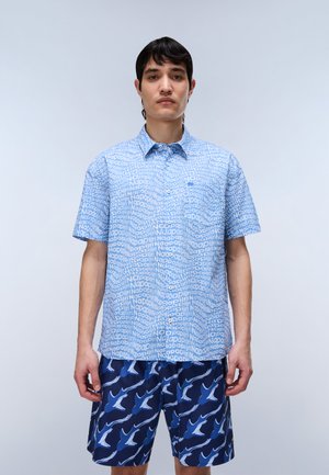 Man met een lichtblauw overhemd met korte mouwen met herhalende tekst "Napapijri" en donkerblauwe korte broek met witte en blauwe haaienpatronen.