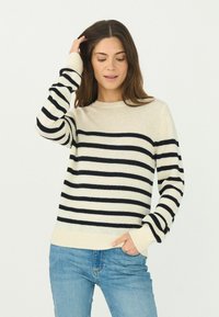 Beige Strickpullover mit marineblauen horizontalen Streifen, Rundhals-Ausschnitt und gerippten Bündchen und Saum. Kombiniert mit blauen Jeans.