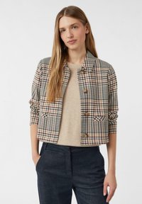 Žena v béžovém svetr pod zapnutou, krátkou kostkovanou bundou s ohrnutými rukávy, v kombinaci s tmavými kalhotami s vysokým pasem, stojící proti bílému pozadí.