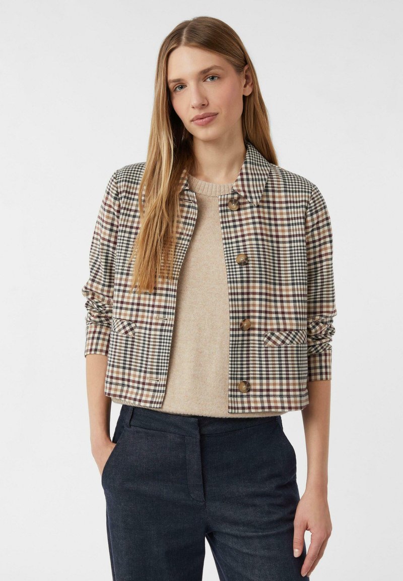 Žena v béžovém svetr pod zapnutou, krátkou kostkovanou bundou s ohrnutými rukávy, v kombinaci s tmavými kalhotami s vysokým pasem, stojící proti bílému pozadí.