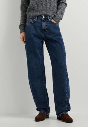 Person trägt lockere dunkelblaue Jeans, einen grauen Strickpullover und braune Wildlederschuhe, steht mit einer Hand in der Tasche.