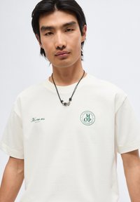 Νέος άνδρας με μαύρα μαλλιά που φοράει λευκό μπλουζάκι με πράσινο κείμενο και μαύρο κολιέ με ασημένιες χάντρες σε ανοιχτό φόντο.