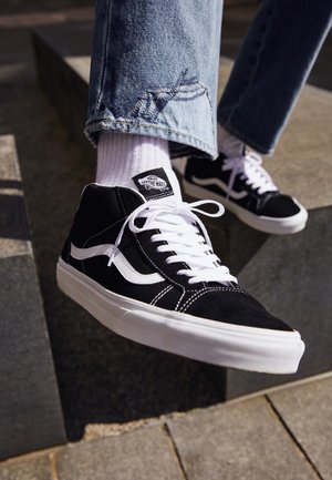 Schwarz-weiße Vans High-Top-Sneaker mit weißen Schnürsenkeln, getragen mit weißen Socken und blauen Jeans, Füße ruhen auf Außentreppe aus Beton.