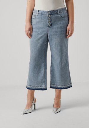 Kvinde iført lyseblå cropped flare jeans med knaplukning og sølvfarvede høje hæle på en ensfarvet baggrund.