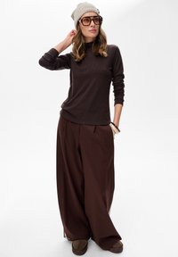 Frau trägt übergroße braune Hosen und ein strukturiertes langärmeliges Oberteil, eine beigefarbene Strickmütze, große Schildkrötensonnenbrillen und Armbänder.