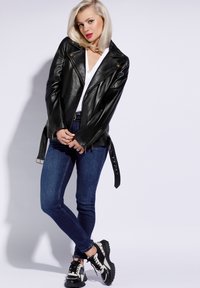 Jachetă de motocicletă din piele neagră cu detalii aurii, asortată cu un top alb și blugi skinny albaștri. Încălțămintea are un design cu model în negru și alb.