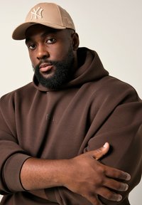 Uomo con barba che indossa un cappellino beige e una felpa marrone, braccia incrociate sul petto, sfondo chiaro neutro.