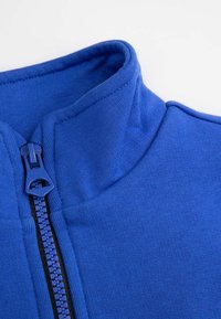 Niebieska polarowa bluzka na zamek błyskawiczny z wysokim kołnierzem, z plastikowym zamkiem i gładkim, teksturowanym materiałem. Prosty design bez wzorów czy akcentów.