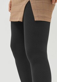Leggings texturizados negros se combinan con una blusa de cuadros marrón, que cuenta con una abertura lateral para mayor movilidad. La tela tiene un acabado suave.