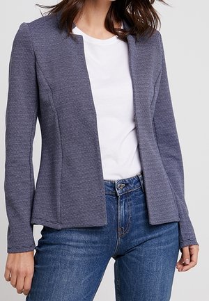 Femme portant un blazer gris texturé ajusté à ouverture frontale sur un t-shirt blanc et un jean bleu, debout devant un fond clair uni.