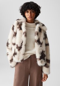Nepluizijakke in crème met bruine abstracte patronen, met een fluffy kraag en een textuurafwerking, gecombineerd met een lichte gebreide trui en bruine broeken.