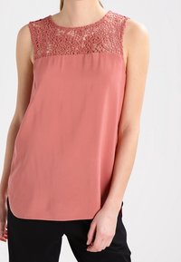 Haut sans manches couleur pêche avec un empiècement en dentelle et un tissu lisse, doté d'un col arrondi et d'une coupe décontractée, associé à un pantalon foncé.