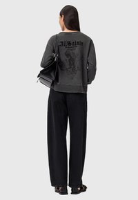 Donna con lunghi capelli scuri che indossa un maglione nero sbiadito con stampa di scheletro, jeans neri larghi, borsa nera e sandali neri, di spalle.