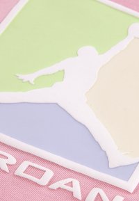 Λαστιχένιο έμβλημα με λευκή σιλουέτα ενός πηδώντας ατόμου· φόντο σε παστέλ πράσινο, μπλε και ροζ· κείμενο "JORDAN" σε λευκό στο κάτω μέρος.