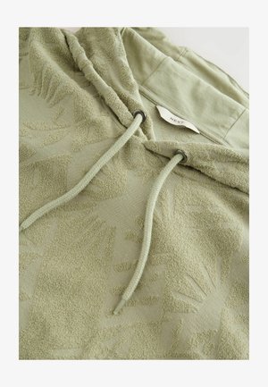 Világos zöld kapucnis pulóver, texturált, emelt levélmintával. Felszereltsége között szerepel egy fémvégekkel ellátott zsinór, valamint egy címke a gallér belsejében.