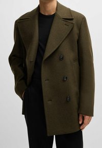 Olivgrön ull dubbelknäppt sjömansjacka med breda slag, fyra svarta knappar och sidfickor. Bärs över en svart skjorta.