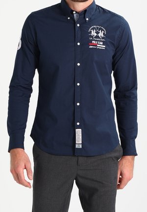 Mann in navyfarbigem Hemd mit La Martina Polo Team-Logo auf der Brust und grauen Hosen, der mit entspannten Armen an den Seiten steht.