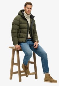 Chaqueta acolchada verde olivo con capucha, detalle de cremallera negra y bolsillos laterales, combinada con un suéter de cuello alto crema, jeans azul y botas marrones.