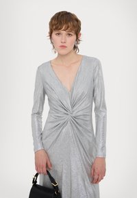 Ασημένιο φόρεμα με μακριά μανίκια, βαθύ V-ντεκολτέ και αναστραμμένη λεπτομέρεια στον κορσέ. Διαθέτει υφή μεταλλικού υφάσματος και πλατιά σιλουέτα.