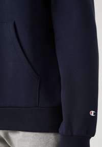 Navy krāsas sporta krekls ar kangaru kabatu, ribinātām piedurknēm un apakšmalu, kā arī ar mazu logo uz kreisās piedurknes. Mīksta, gluda auduma tekstūra.