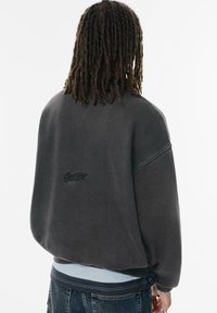 Ciemnoszary, oversize'owy sweter z okrągłym dekoltem, obniżonymi ramionami oraz delikatnie haftowanym logo na plecach. Miękka, teksturowana tkanina.