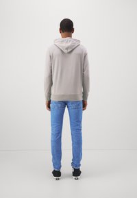 Šedá mikina s kapucí, měkká tkanina, přiléhavý střih. V kombinaci s modrými džínami, slim fit. Tmavé boty s bílými akcenty. Pohled zezadu.