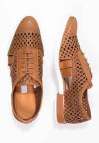 Scarpe in pelle marrone perforate con suola bianca, caratterizzate da un design con lacci e modelli intagliati per traspirabilità e texture.