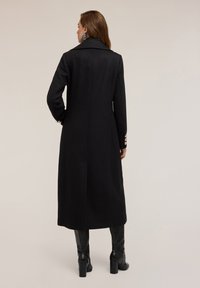 Cappotto lungo nero in tessuto strutturato, con silhouette dritta, grande colletto e dettagli con bottoni sui polsini. Texture liscia, design minimale.