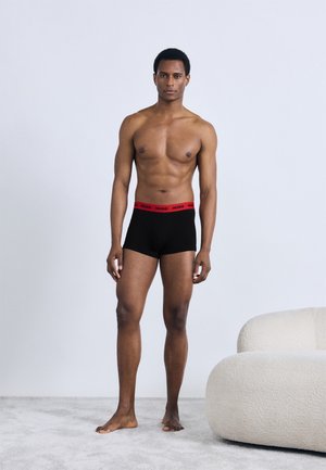 Boxers noirs avec une ceinture élastique rouge portant le logo "HUGO", portés par un mannequin, présentés sur un fond neutre avec un sol à texture douce.