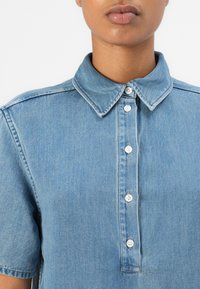 Lys blå denimskjorte med krage, korte ermer og en frontpanel med hvite knapper.