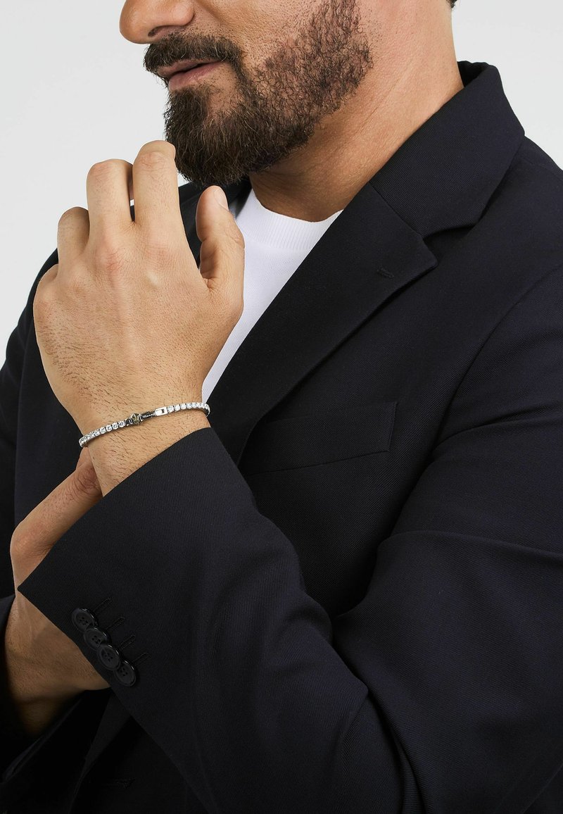 Bracciale in argento con dettaglio di chiusura, indossato su un polso nudo, abbinato a un blazer nero. La trama del tessuto è liscia e lucida.