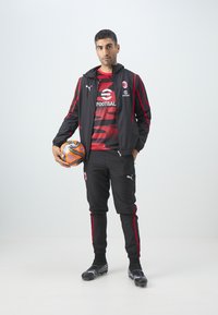 Chándal atlético negro con acentos rojos, que cuenta con una camiseta estampada y pantalones a juego. El modelo sostiene un balón de fútbol naranja y negro, vistiendo botas de futbol.