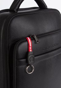 Musta teksturoitu synteettinen laukku vetoketjulla, punaisella ja valkoisella vetonauhalla sekä pyöreällä metallirenkaalla sivussa.